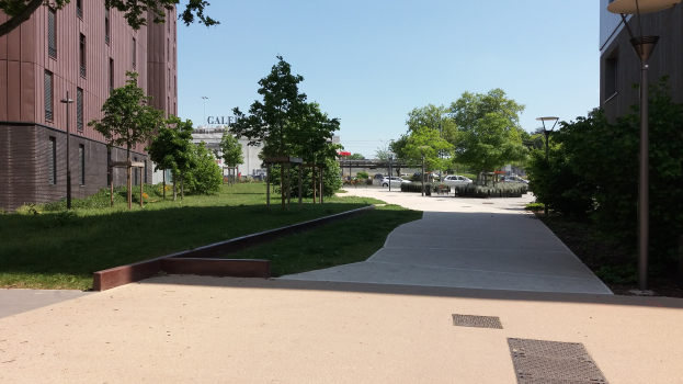 Ein gepflasterter Weg in einem Park mit Bäumen und Gras, flankiert von hohen Gebäuden, mit einer Straße mit Fahrzeugen, Laternen und Pfählen, der zu einer Brücke unter einem klaren blauen Himmel führt.