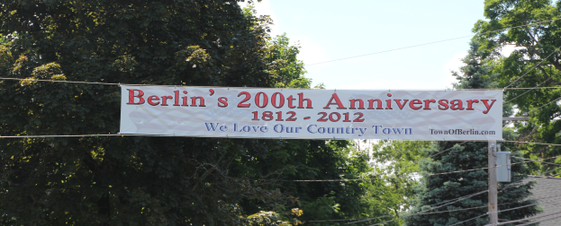 Ein Banner mit der Aufschrift 'Berliner Jubiläum' hängt an einem Draht in der Mitte, umgeben von Bäumen und einem Haus auf der rechten Seite, mit einem sichtbaren Himmel.