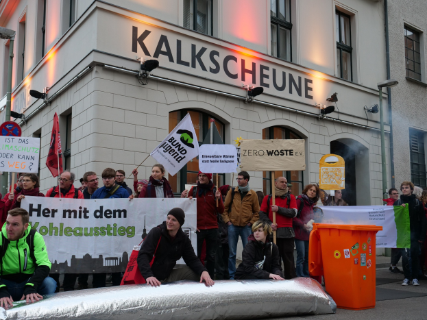 Eine Gruppe von Menschen mit Schildern und Plakaten vor einem Gebäude bei einer Demonstration in Deutschland, mit zwei Menschen auf einem Gegenstand sitzend im Vordergrund und einem Mölltonnen auf der rechten Seite.