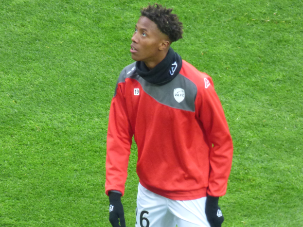 Ein junger Mann in einer roten Jacke und weißen Shorts steht auf einem Fußballfeld und schaut entschlossen nach rechts, während er Baseballhandschuhe trägt.