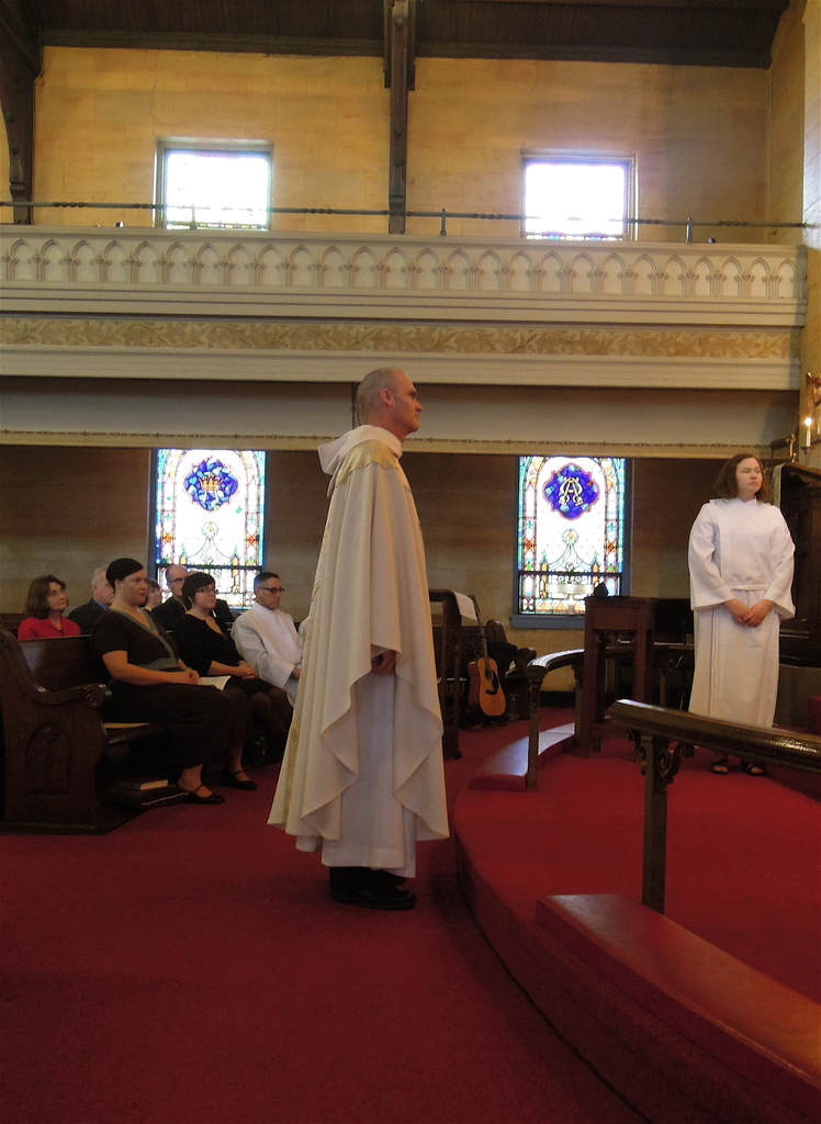 Eine Frau und ein Mann in weißen Kleidern stehen in der Mitte einer Kirche, mit einem roten Teppich zu ihren Füßen, einem hölzernen Geländer und einer Kerze hinter der Frau, Menschen, die auf Bänken links sitzen, Musikinstrumente im Hintergrund und Geländern, Fenstern und Wänden im gesamten Bild.