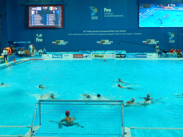 Eine Gruppe von Menschen, die Wasserball in einem großen Pool mit einem Netz in der Mitte spielen, Zuschauer auf der linken Seite und eine Wand mit einem Bildschirm im Hintergrund bei den FINA-Weltmeisterschaften 2016 in Kazan.