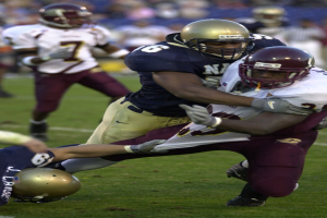 Zwei Füssballspieler in Helmen und Uniformen stoßen während eines Spiels zusammen, mit anderen Spielern und Zuschauern im Hintergrund.