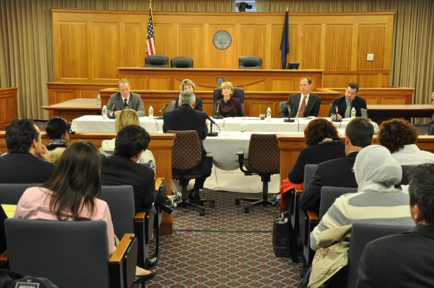 Ein Versammlungsraum mit einer Gruppe von Menschen auf Stühlen, zwei ausländischen Flaggen, einem orangen Schreibtisch mit Wasserflaschen und einem Mikrofon.