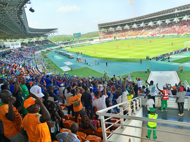 Eine große Menschenmenge in einem Stadion bei einem Fuballspiel, mit einem Schuppen mit Lichtern auf der linken Seite und Hügel unter einem klaren blauen Himmel im Hintergrund.