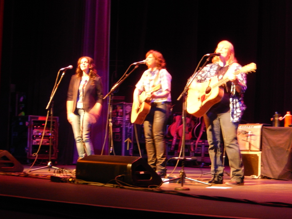 Drei Frauen treten auf der Bühne eines Musikkonzerts auf, jede singt in ein Mikrofon, während sie eine Gitarre spielen, mit einem Tisch mit Wasserflaschen und zusätzlichen Gitarren dahinter.