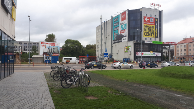 Stadtstraße mit Gebäuden, Straßenlaternen, fahrenden Kraftfahrzeugen, Fußgängern auf dem Gehweg, parkenden Fahrrädern, Werbetafeln, Bäumen und einem bewölkten Himmel.