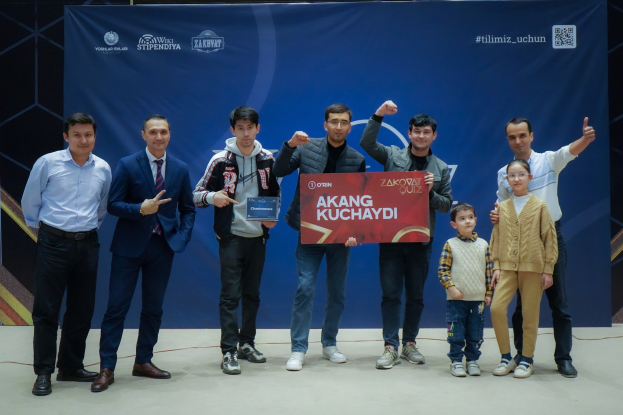Gruppe von Menschen auf einer Bühne mit einer Tafel, auf der "Akang Kuchaydi - Zakovat Quiz" steht, im Hintergrund ein Banner und Lichter.