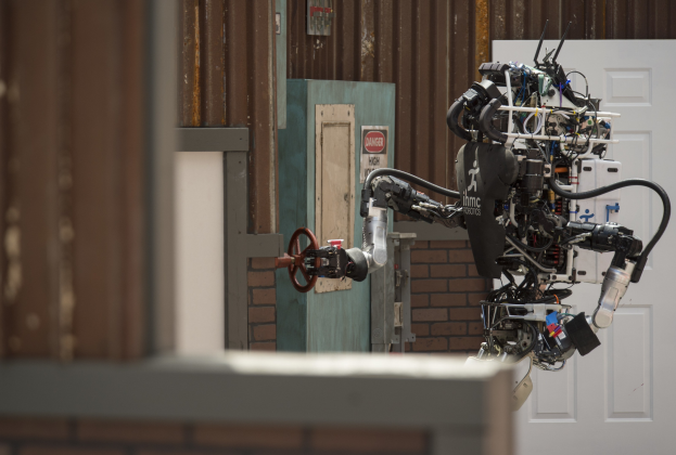 Ein humanoider Roboter mit ausgestreckten Armen und geneigtem Kopf steht vor einem Gebäude mit einer Tür und einer Wand im Hintergrund, seine großen runden Augen und leicht geöffnete Mund deuten darauf hin, dass er gleich sprechen wird.