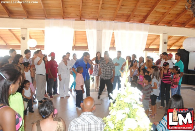 Eine Gruppe von Menschen mit einer Pflanze und einem Logo unten, Vorhängen und einem Lautsprecher im Hintergrund und Text in der oberen linken Ecke.