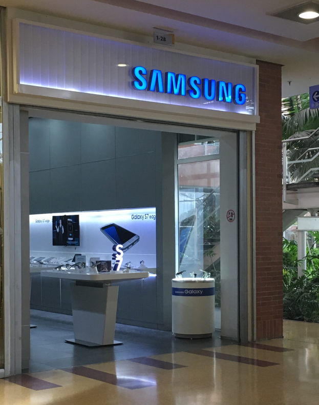Ein Samsung-Geschäft in einem Einkaufszentrum mit einem Namensschild an der Wand, einem Tisch mit verschiedenen Gegenständen, einem Schild, einer Gruppe von Pflanzen, einer Treppe und einer Decke mit Leuchten.