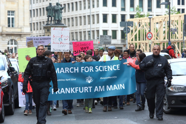 Eine Gruppe von Menschen marschiert auf einer Straße und hält ein "March for Science Frankfurt am Main"-Schild hoch, mit Autos, Gebäuden, Statuen, Laternenpfählen, Schildern und Bäumen im Hintergrund.