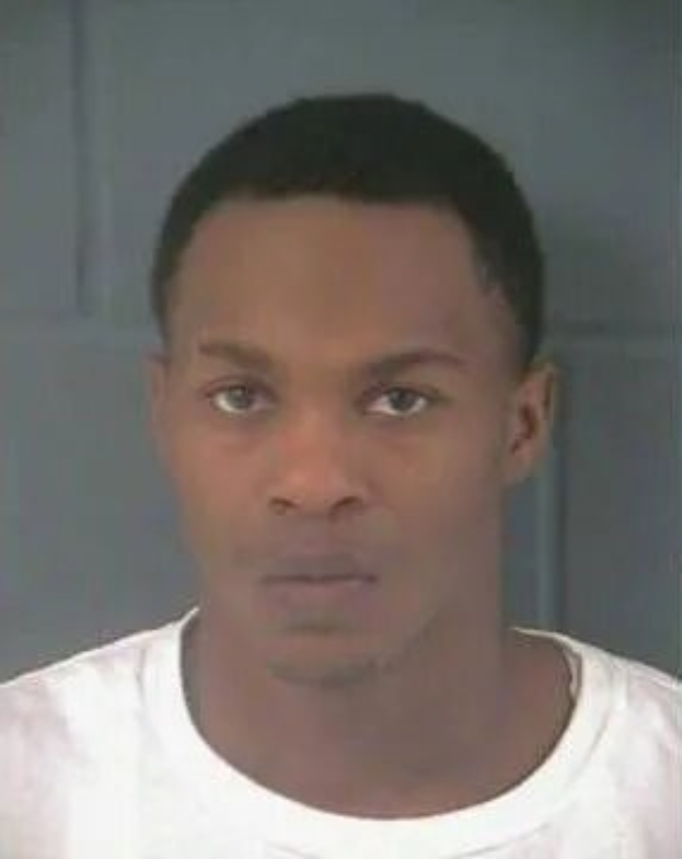 Ein Bild eines Mannes in einem weißen T-Shirt vor einer Wand, der verdächtigt wird, in einen Mordfall verwickelt zu sein.