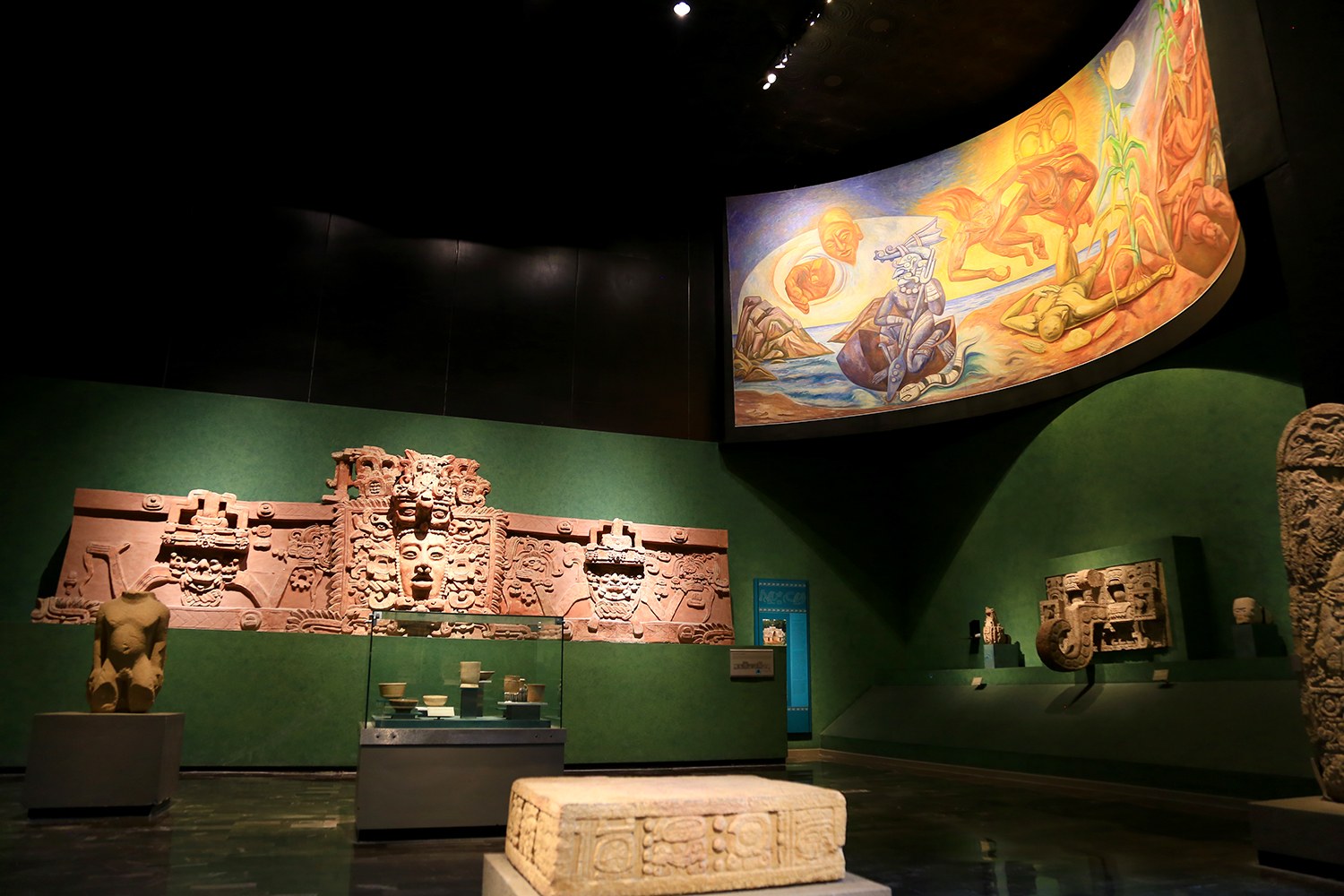 Ein Museumsraum mit verschiedenen Artefakten, darunter Skulpturen, Tischgegenstände, eine Texttafel, eine Wandmalerei und Deckenleuchten im Anthropologie-Museum von Mexiko-Stadt.