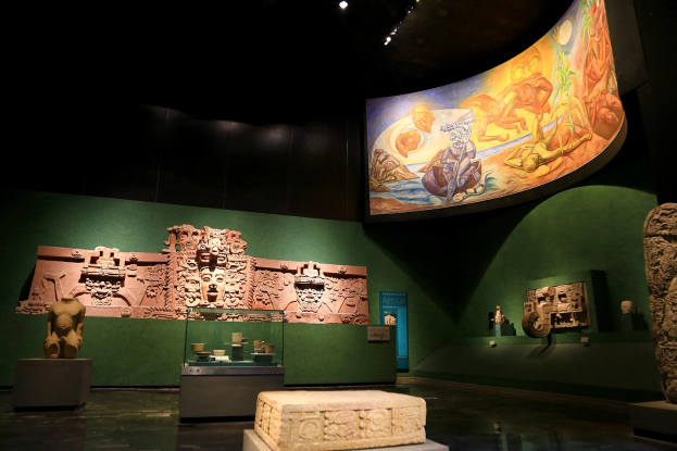 Ein Museumsraum mit verschiedenen Artefakten, darunter Skulpturen, Tischgegenstände, eine Texttafel, eine Wandmalerei und Deckenleuchten im Anthropologie-Museum von Mexiko-Stadt.