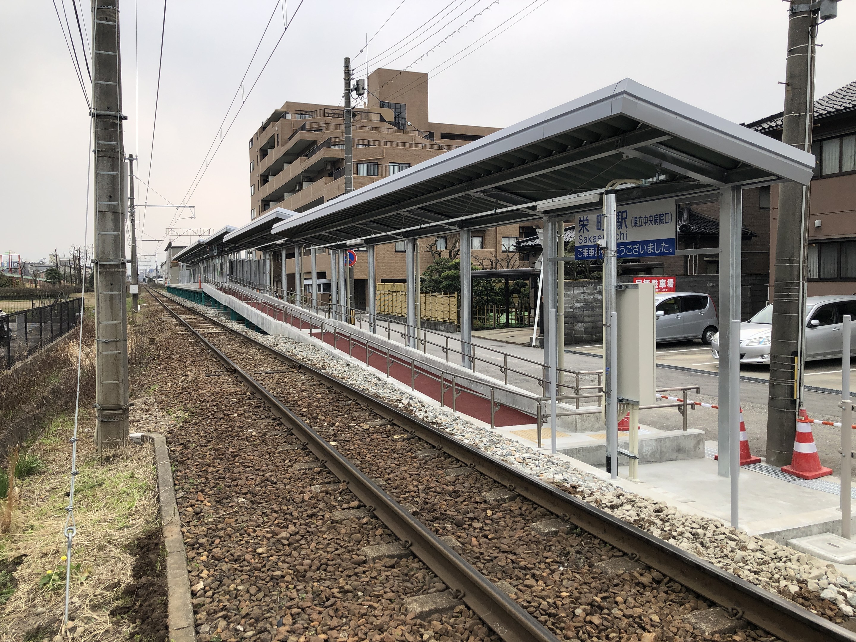 Ein Bahnhof mit einem Zug auf den Schienen, umgeben von Gebäuden, Strommasten, Stromkabeln, Kraftfahrzeugen auf der Straße, Absperrgittern, Informationsschildern, Zäunen, Bäumen und einem klaren blauen Himmel.