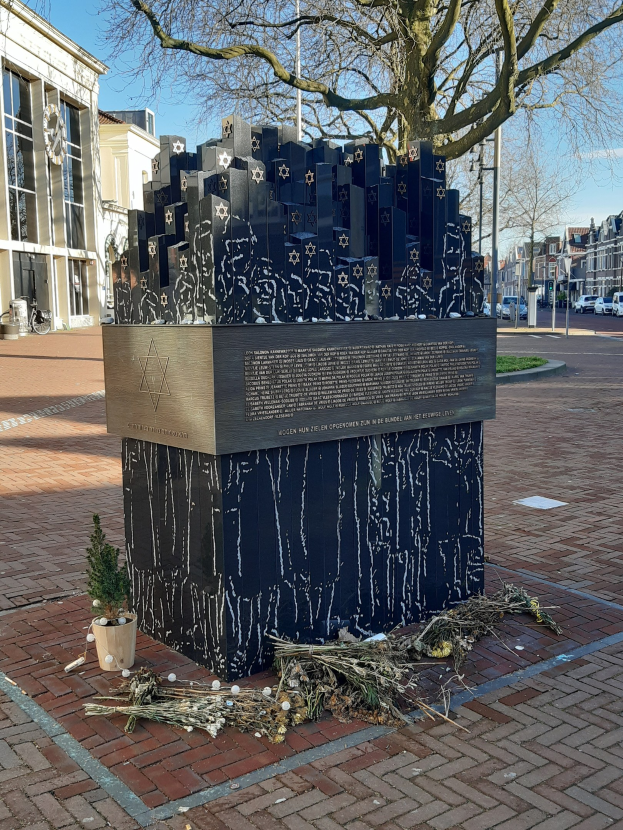 Denkmal in einem Stadtplatz für Holocaust-Opfer mit einem Baum davor, einem Topfgewächs, verstreuten trockenen Blättern und Gebäuden, Fahrzeugen, Pfählen und Bäumen im Hintergrund.