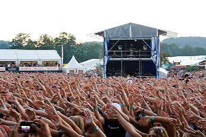 Eine große Menge auf einem Musikfestival mit erhobenen Händen, einige halten Handys, vor einer Bühne mit Könpern, weiße Zelte, Bäume und einen klaren blauen Himmel.