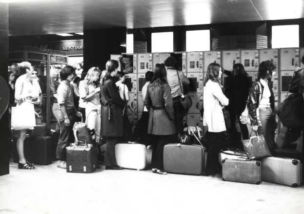 Schwarzes und weißes Foto einer Gruppe von Menschen mit Gepäck, die in einem Flughafen stehen, mit Sicht auf Schließfächer im Hintergrund.