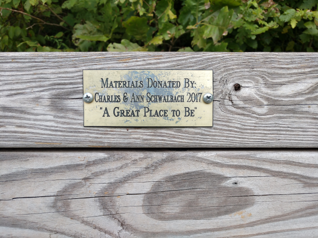 Eine hölzerne Bank mit einer Plakette, auf der steht: "Spende von Charles & Ann Schwabach 2017: Ein toller Ort zu sein" umgeben von üppigen grünen Pflanzen.