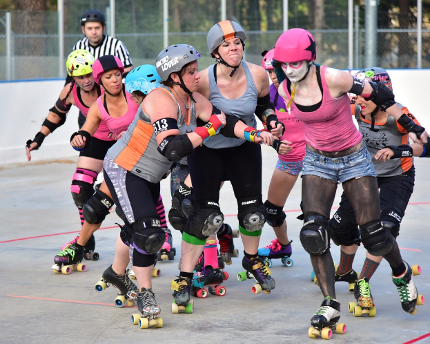 Mehrere Frauen mit Helmen und Schlittschuhen schieben sich gegenseitig auf Kufen, ein Mann steht daneben, ein Metallzaun und Bäume im Hintergrund.