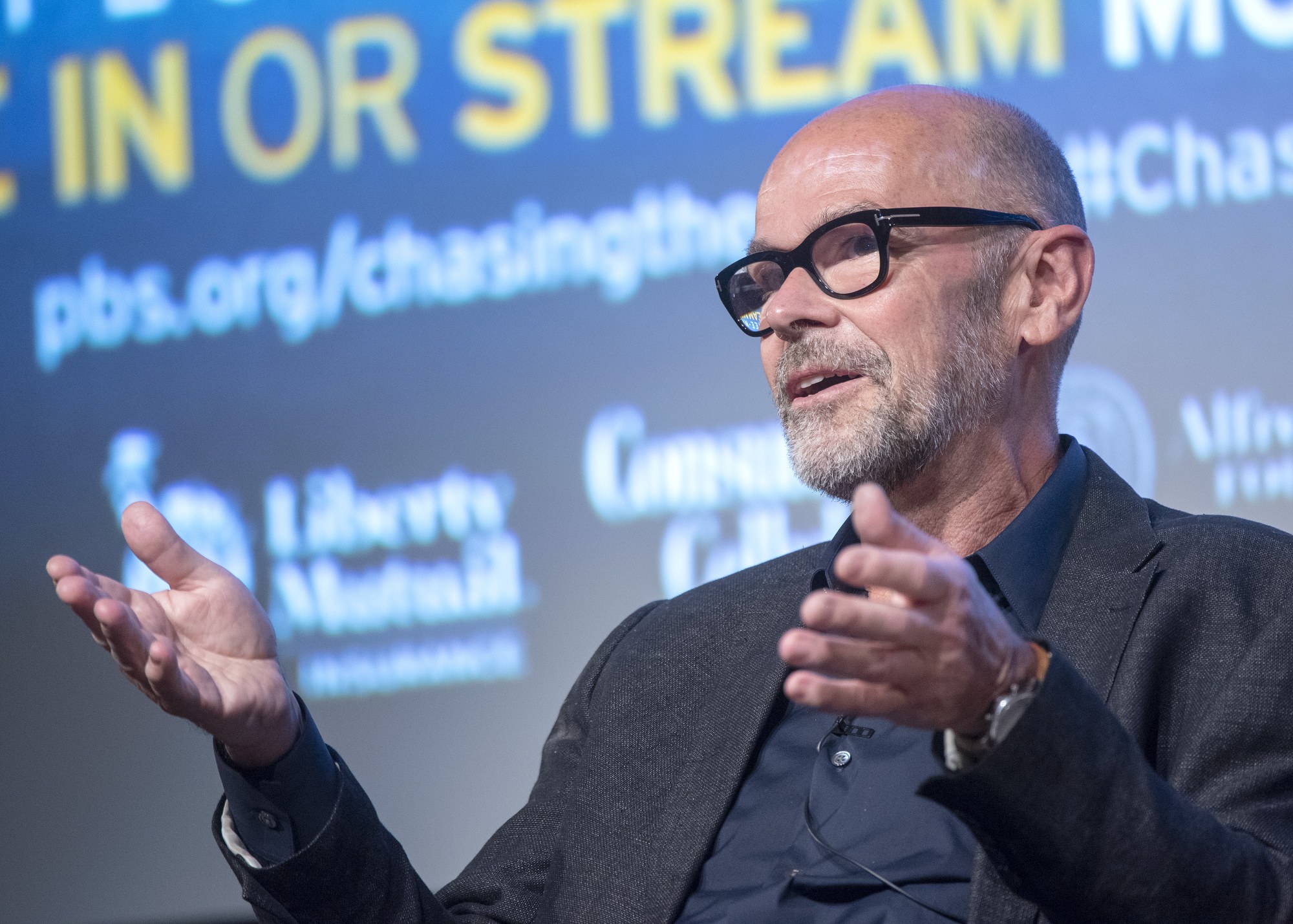 Steve Ballmer, Gründer und CEO von PBS, spricht in ein Mikrofon, während im Hintergrund ein Bildschirm mit Text zu sehen ist.