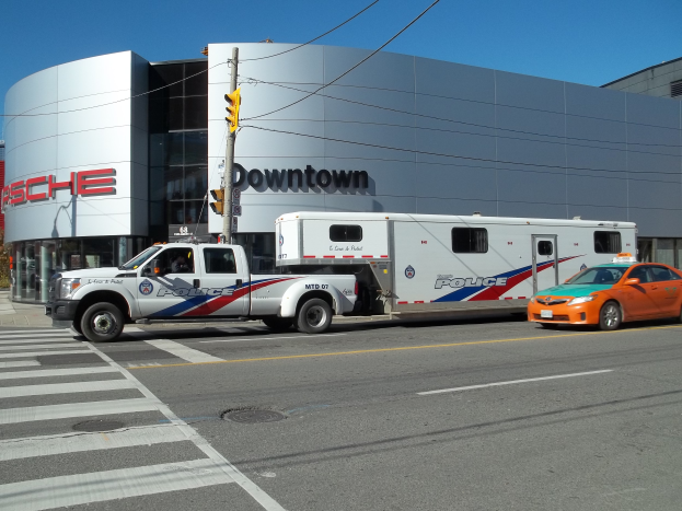Ein vor einem Gebäude parkender Polizeiwagen mit Text, ein Verkehrszeichen mit Stange und Drähten im Vordergrund und Bäume mit klarem blauem Himmel im Hintergrund.