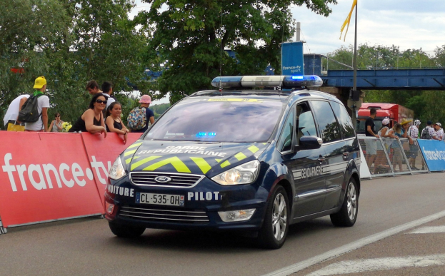 Polizeiauto fährt auf einer Straße neben einer Menschenmenge mit Schildern und Absperrungen im Hintergrund, Bäume, eine Brücke, eine Fahne und einen bewölkten Himmel.