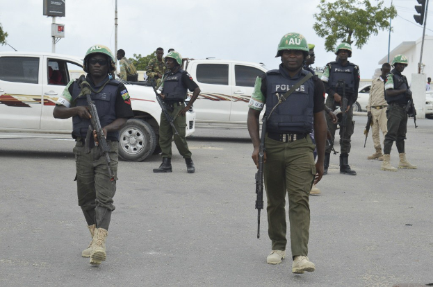 Eine Gruppe von Polizeibeamten in Helmen und bewaffnet mit Gewehren geht eine Straße entlang mit Fahrzeugen, Verkehrszeichen, Bäumen, Gebäuden und einem bewölkten Himmel im Hintergrund.