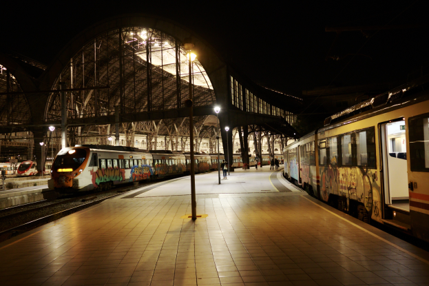 Ein Bahnhof in der Nacht mit zwei geparkten Zügen, einigen Menschen auf dem Bahnsteig und Laternen, die das Gebiet beleuchten, vor einem dunklen Hintergrund.
