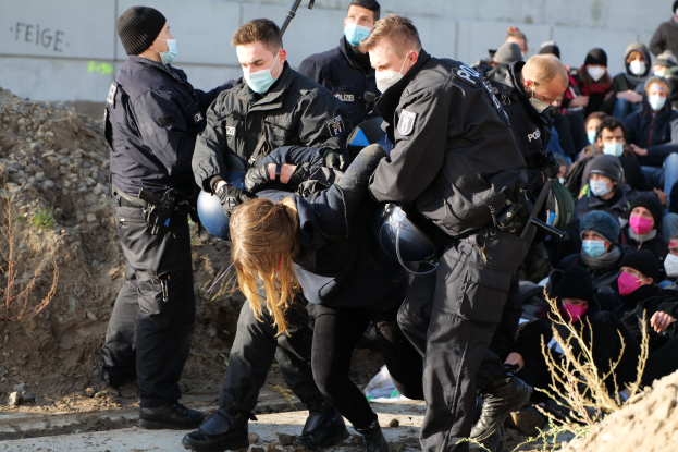 Gruppe von maskierten Menschen in Mützen, einige mit Waffen, die eine von der Polizei begleitete Frau umgeben, vor einer Wand mit Text und Pflanzen in einer städtischen Umgebung, was auf eine Protestaktion gegen Regierungssparmaßnahmen hinweist.