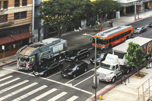 Eine belebte Stadtstraße mit Autos, Bussen, Lastwagen, Bäumen, Laternenmasten, Gebäuden, Geländern, Treppen, Menschen auf dem Gehweg und Schildern.