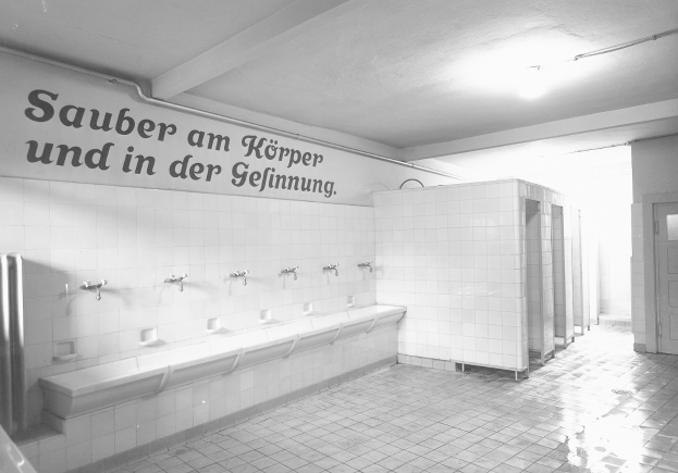 Schwarz-weiß-Foto einer öffentlichen Toilette mit Fliesenboden, einer textbedeckten Wand, mehreren Waschbecken, einer Tür und einer deckenmontierten Lampe.