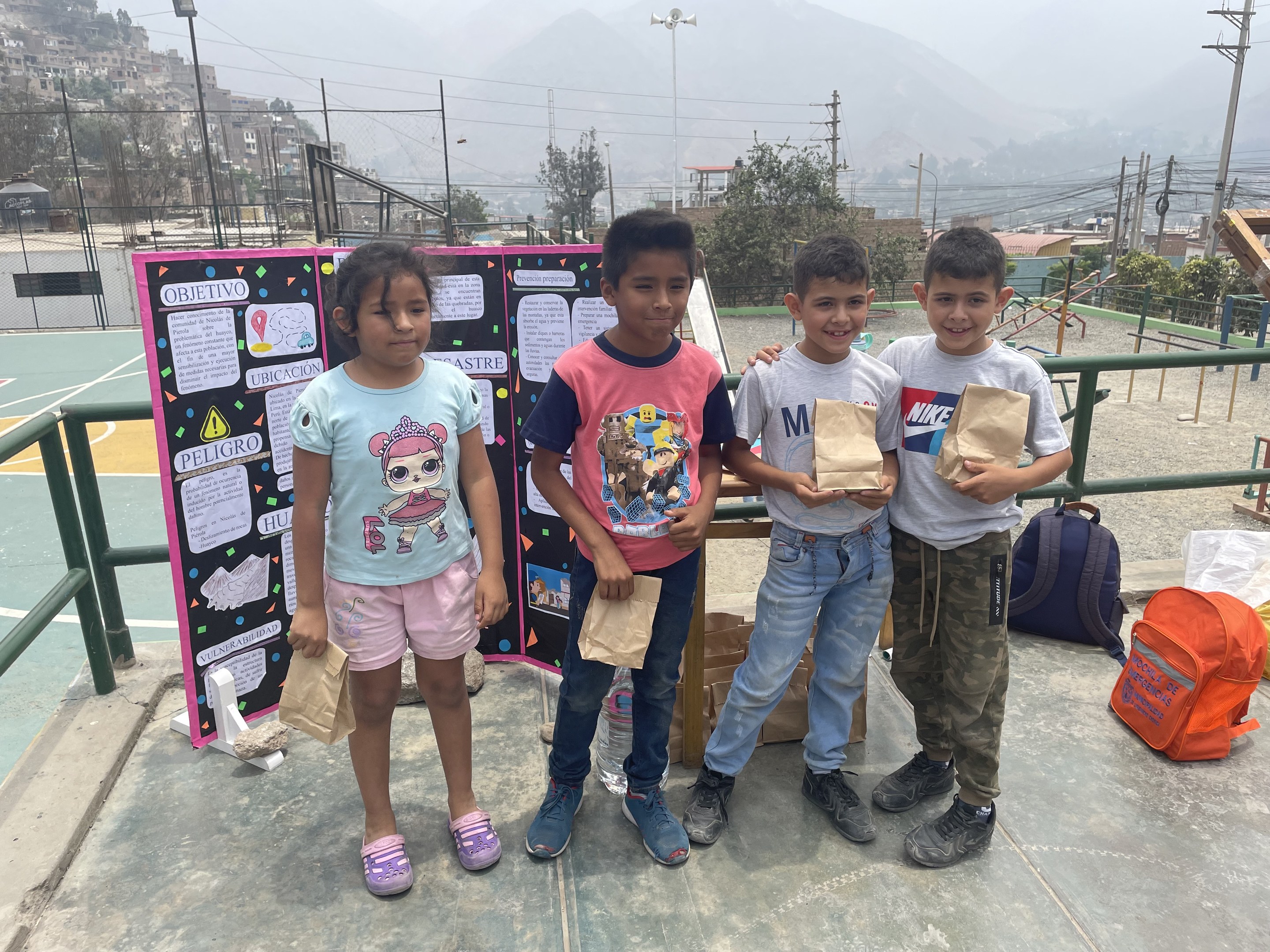 Gruppe von Kindern auf einem Basketballfeld mit Papiertüten in den Händen, Gegenstände auf der rechten Seite und eine Tafel mit Text auf der linken Seite, vor einem Hintergrund aus Zaun, Pfosten, Straßenlaternen, Bäumen, Gebäuden, Hügeln und Himmel.