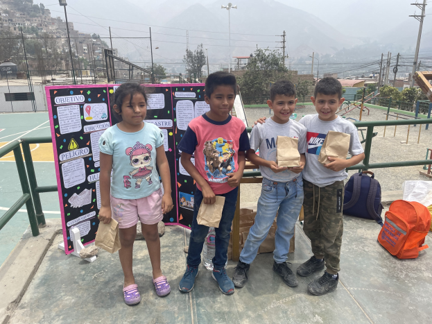 Gruppe von Kindern auf einem Basketballfeld mit Papiertüten in den Händen, Gegenstände auf der rechten Seite und eine Tafel mit Text auf der linken Seite, vor einem Hintergrund aus Zaun, Pfosten, Straßenlaternen, Bäumen, Gebäuden, Hügeln und Himmel.