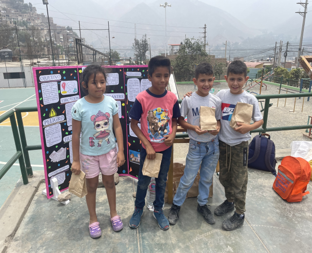 Gruppe von Kindern auf einem Basketballfeld mit Papiertüten in den Händen, Gegenstände auf der rechten Seite und eine Tafel mit Text auf der linken Seite, vor einem Hintergrund aus Zaun, Pfosten, Straßenlaternen, Bäumen, Gebäuden, Hügeln und Himmel.