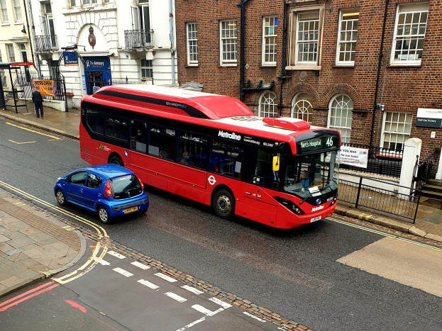 Roter Doppeldeckerbus fährt auf einer Straße neben einem blauen Auto, mit Füßgängern auf einem Gehweg und Gebäuden im Hintergrund.