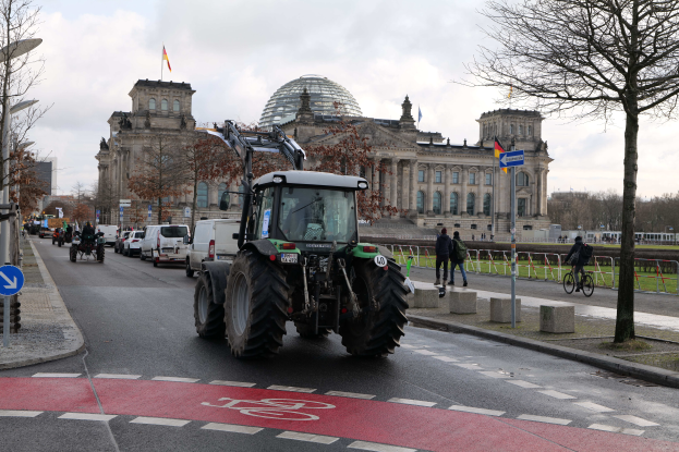 Ein Traktor fährt eine Straße vor dem Reichstaggebäude in Berlin, Deutschland, entlang, mit Bäumen, Laternen und Schildern an der Straße und Menschen, die zu Fuß und mit Fahrrädern auf dem Gehweg unterwegs sind, unter einem bewölkten Himmel mit Fahnen am Gebäude.