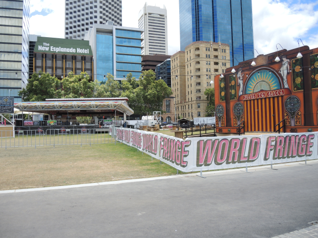 Eine Karnevalsattraktion ist in einer Stadtstraße aufgebaut, umgeben von hohen Gebäuden, Bäumen und einem bewölkten Himmel, mit einer Straße, einem Geländer und einer Banner mit Text im Vordergrund.