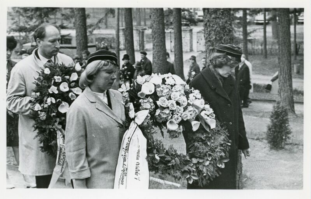 Eine Gruppe von Menschen hält Blumensträuße in den Händen und steht zusammen bei einer Holocaust-Gedenkfeier, mit Bäumen und Gebäuden im schwarz-weißen Hintergrund.