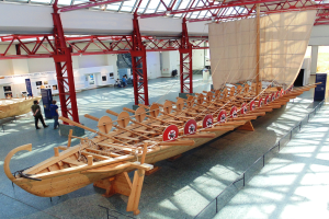 Ein großes hölzernes Wikingerschiff in einem Museum ausgestellt, umgeben von Menschen, Metallstangen, einem Zaun, einer Fahne, einer Wand und einer Decke mit Lampen.