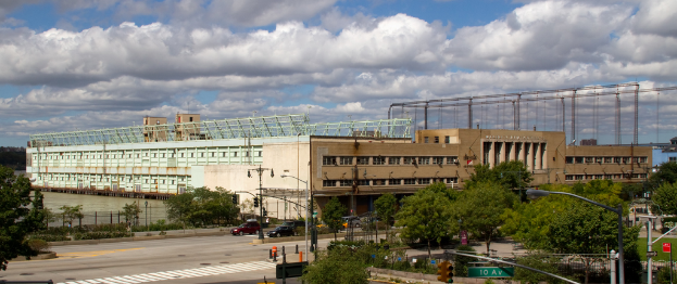 Eine Straße mit Fahrzeugen, Bäumen und Straßenlaternen entlang eines Wasserlaufs mit einem großen Gebäude im Hintergrund.