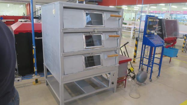 Ein Labor mit elektrischen Geräten und Metallregalen im Vordergrund, einer Person links und einer Glaswand mit ein paar Personen dahinter rechts.