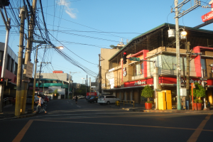 Eine Stadtstraßenszene mit Autos, Fußgängern, Strommasten, Verkehrszeichen, Gebäuden mit Schildern, Topfpflanzen und einem bewölkten Himmel.