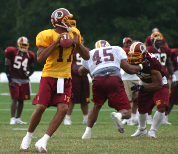 Eine Gruppe von American-Football-Spielern in Trikots, Helmen und weißen Schuhen auf einem grasbewachsenen Feld mit Bäumen im Hintergrund.