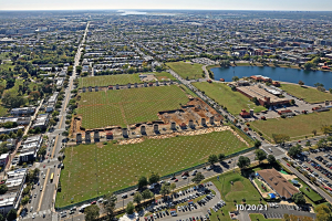 Luftaufnahme einer Stadt mit Gebäuden, Bäumen, Gras, Fahrzeugen, Wasser und Wolken, die einen großen zentralen Platz zeigt, der als Parkgebiet vorgeschlagen ist.
