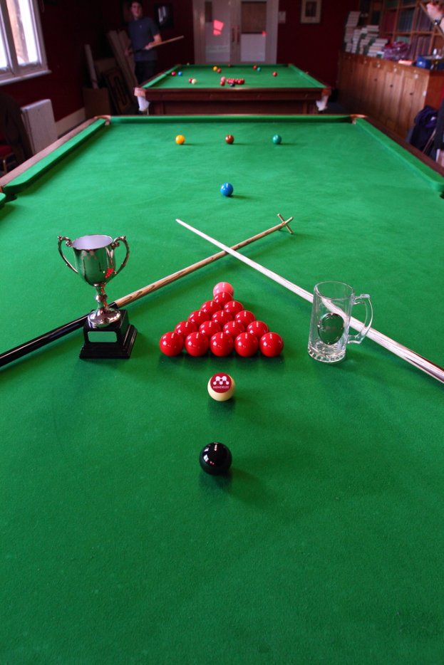 Ein Pokal, ein Glasbecher und mehrere Kugeln sind auf einem Billardtisch angeordnet.