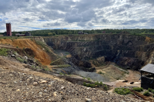 Großer Tagebau auf einem Schmutzfeld umgeben von Bäumen, Gebäuden und einem Schuppen, mit einem bewölkten Himmel und einem mit Steinen und Pflanzen bedeckten Boden.