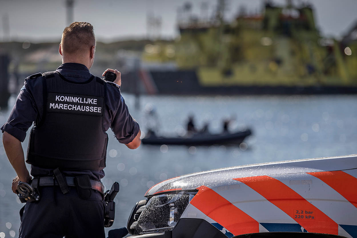 Ein Polizist in Uniform steht neben einem Polizeiauto in der Nähe eines Gewässers und hält ein Funkgerät in der Hand, im Hintergrund ist ein Boot mit ein paar Menschen auf dem Wasser zu sehen und der Himmel darüber.
