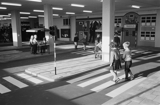 Schwarz-weiß-Foto von Kindern, die eine Straße an einer Fußgängerampel überqueren, mit Fußgängern in der Nähe, einem Verkehrssignalmast im Vordergrund und strukturellen Säulen und Leuchten im Hintergrund.